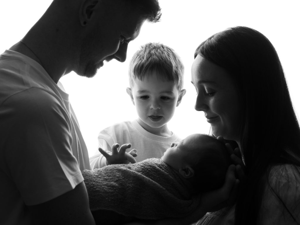 Black and white portrait of two parents and a young child gazing tenderly at a swaddled newborn baby