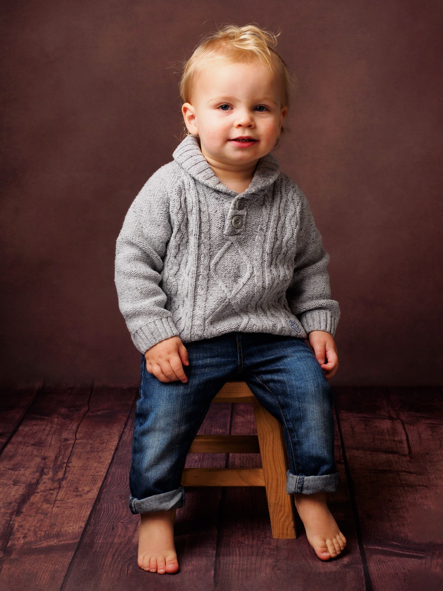 Little boy in a grey cable knit jumper sitting on a wooden stool