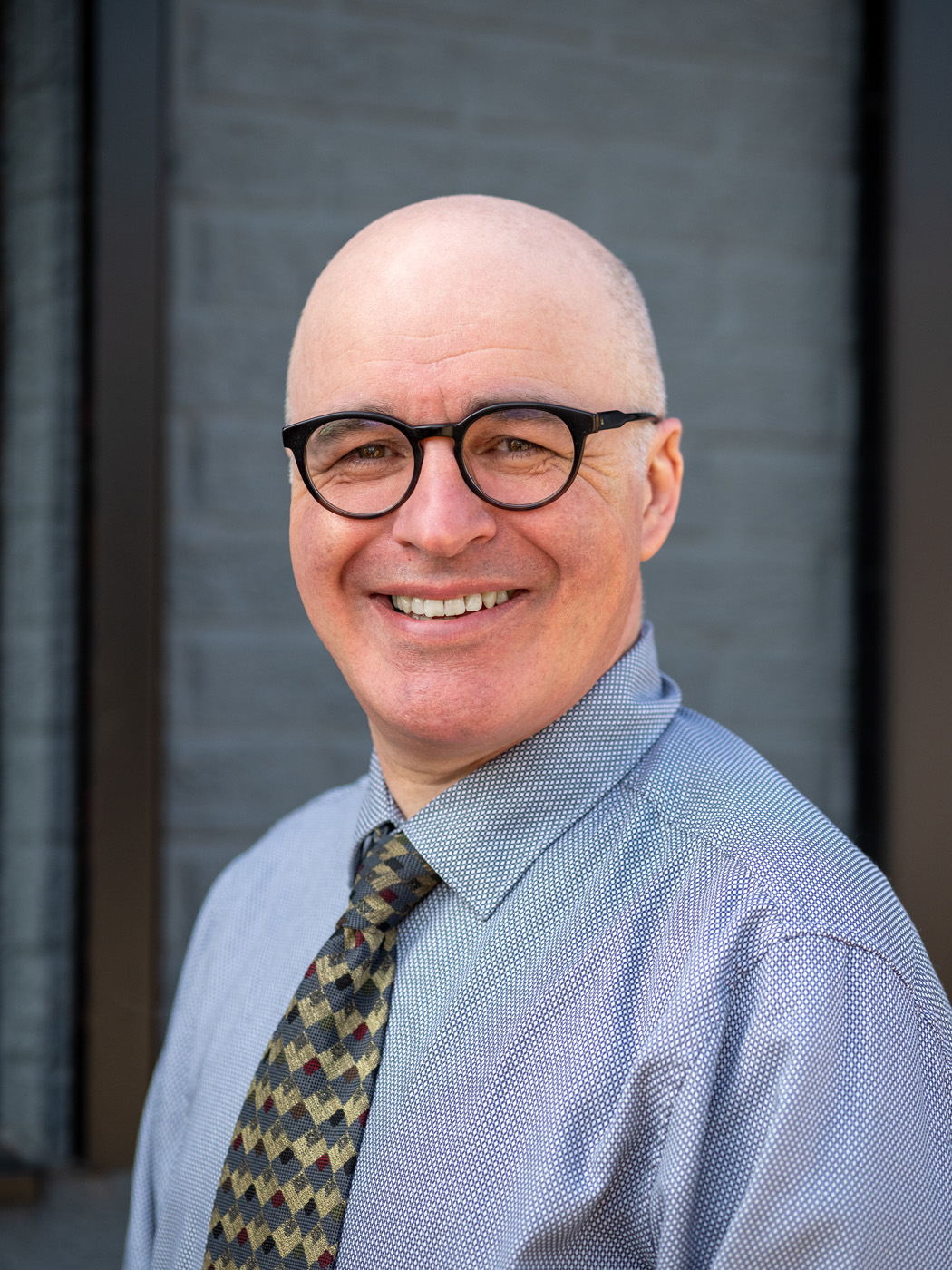 Smiling business headshot of a man in round glasses and a patterned tie against a grey wall