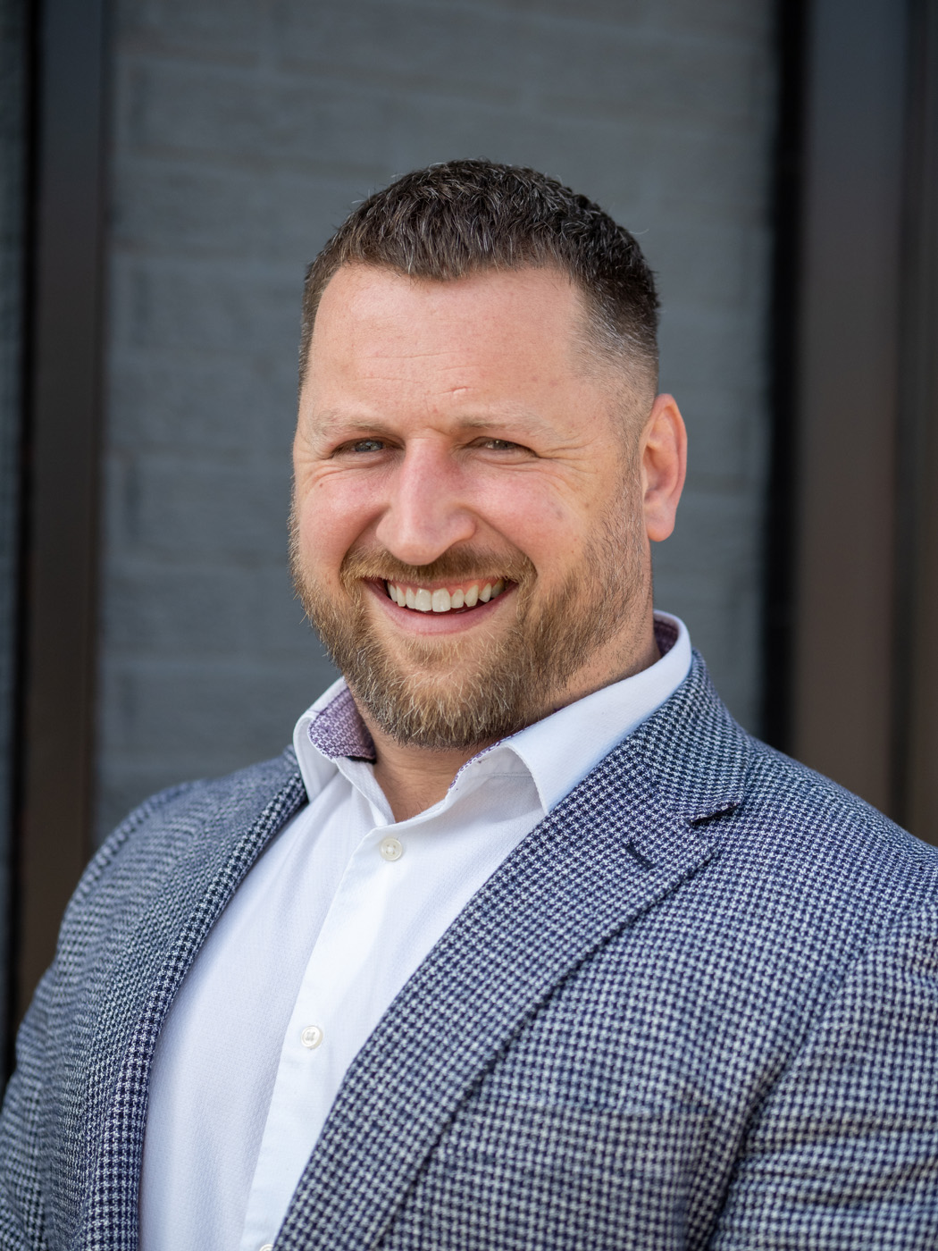 Smiling business headshot of a bearded man in a houndstooth blazer against a grey wall