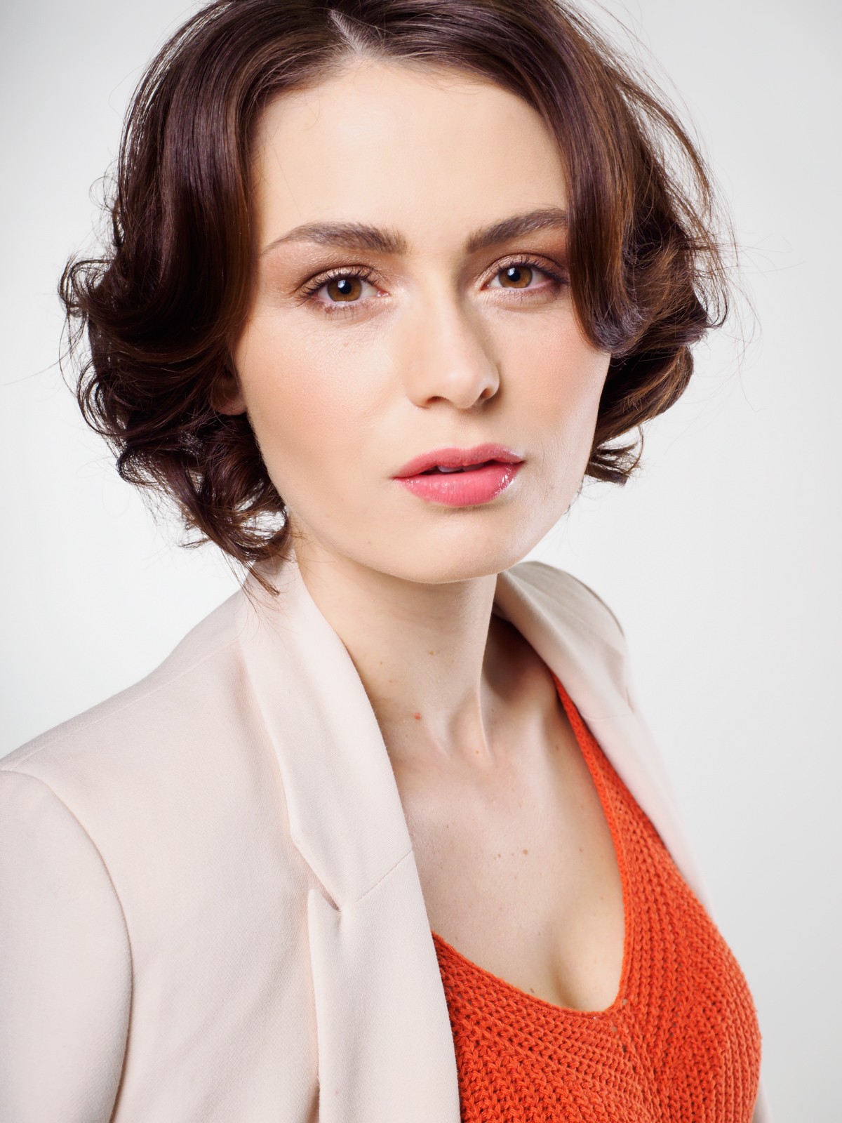 Actor headshot of a woman with short dark curls in a pale blazer and orange top