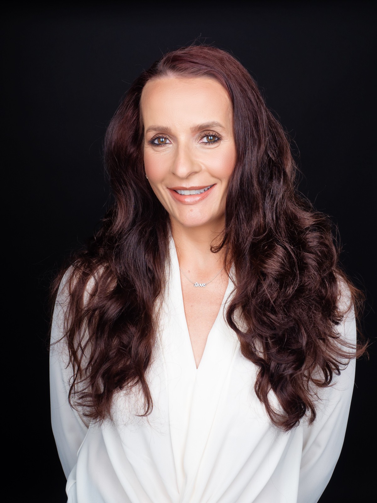 Smiling business headshot of a woman with long dark curls in a white blouse on black